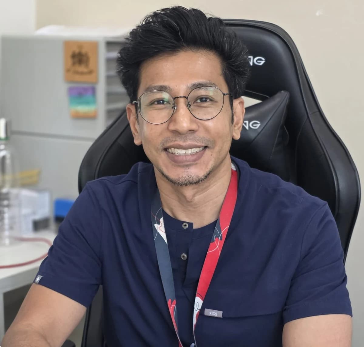 Dr Sulaiman Bin Yusof seated at his consulting room desk in navy scrubs and round glasses, facing the camera and smiling.