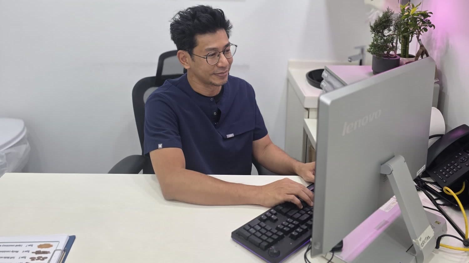 Dr Sulaiman Bin Yusof in navy scrubs seated at his clinic desk, one hand on the mouse, looking at the monitor in front of him.
