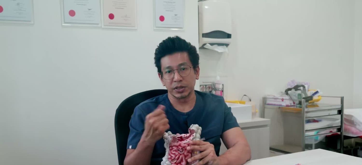 Dr Sulaiman Bin Yusof at his clinic desk, holding an anatomical model of the large and small intestine in one hand and gesturing with the other as he explains.