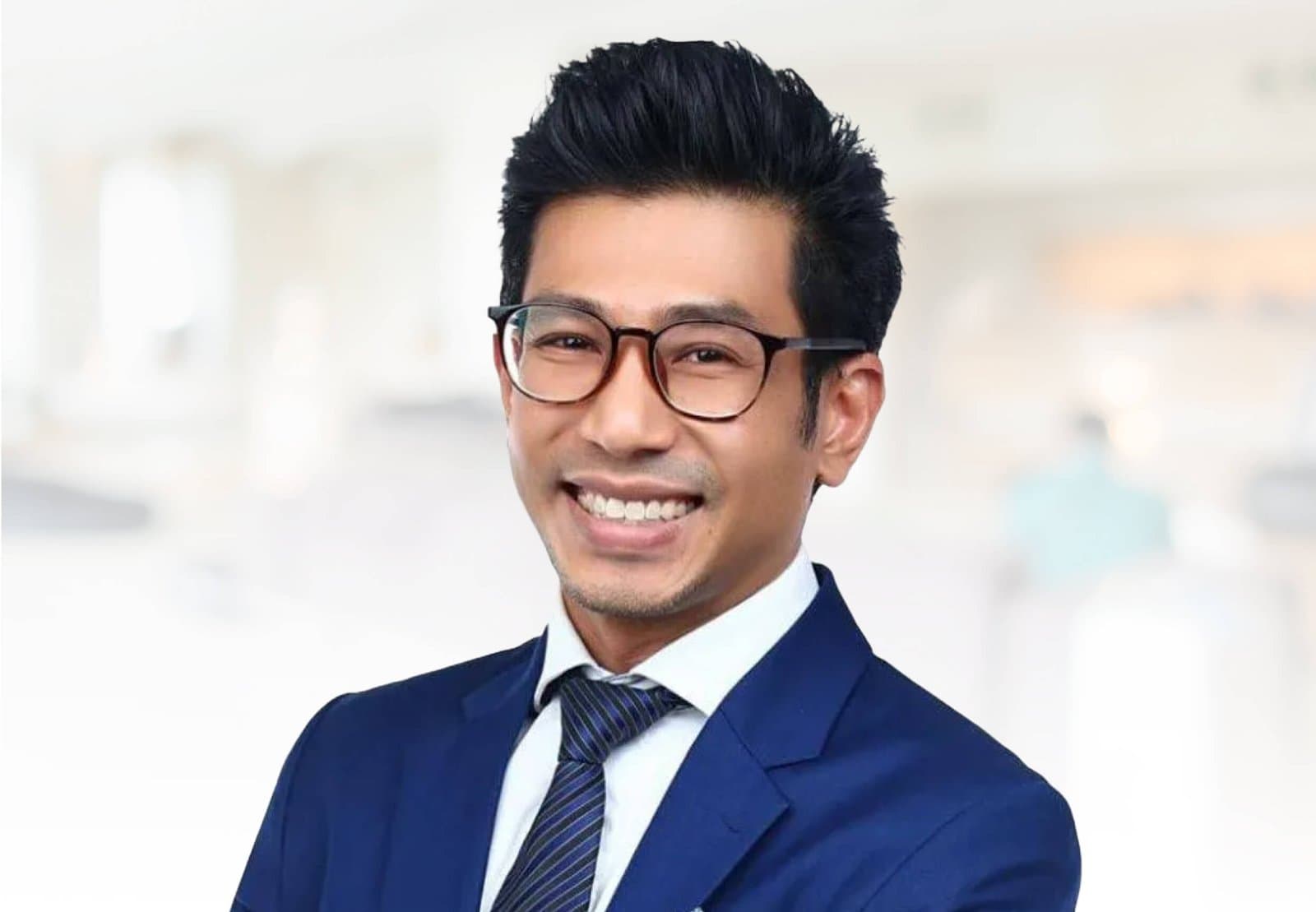Dr Sulaiman Bin Yusof in a navy suit and striped tie, wearing dark-framed round glasses, photographed against a softly blurred clinic interior.