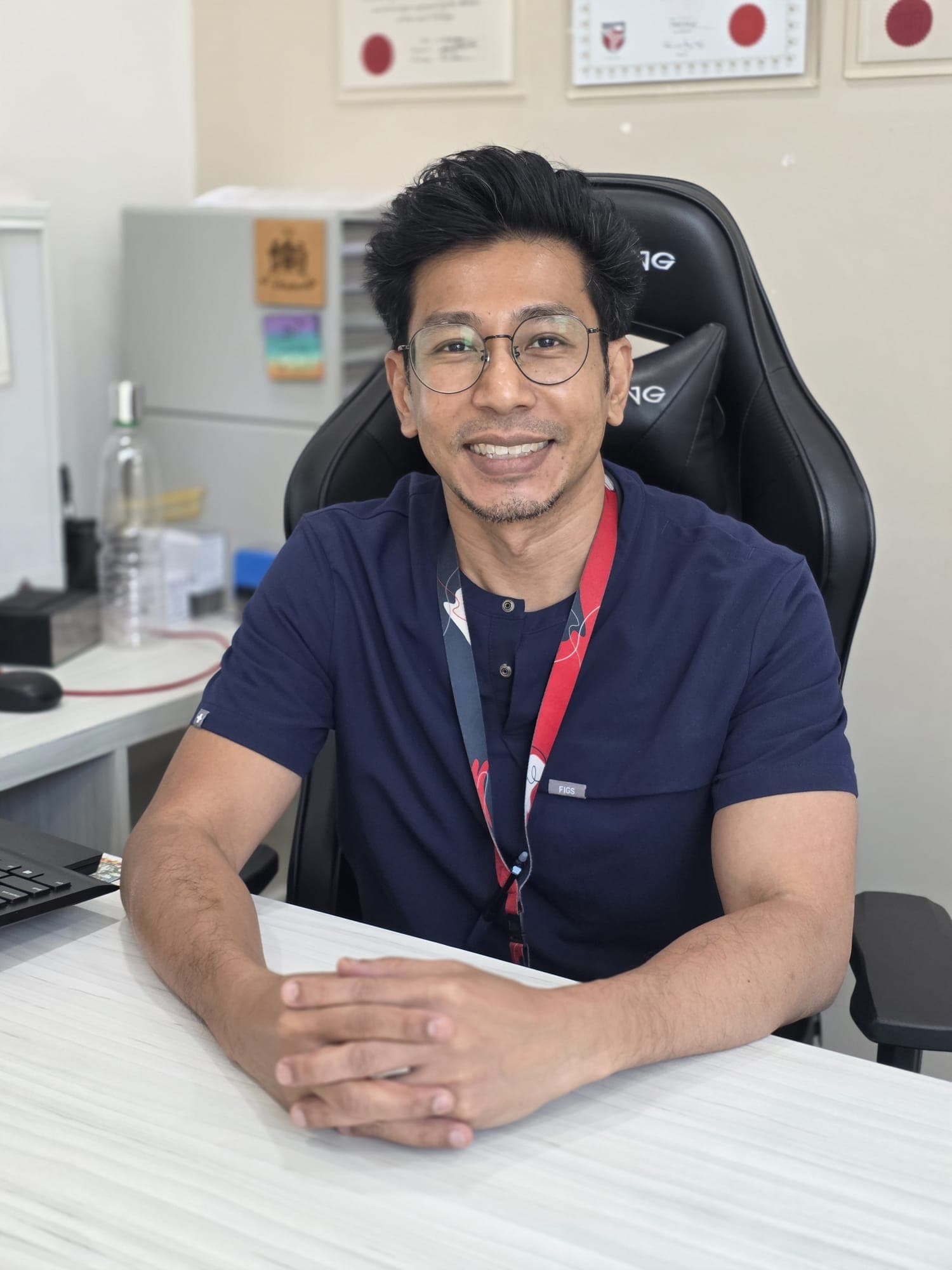 Dr Sulaiman Bin Yusof in navy scrubs, seated at his consult-room desk with his hands clasped in front of him, smiling towards the camera.