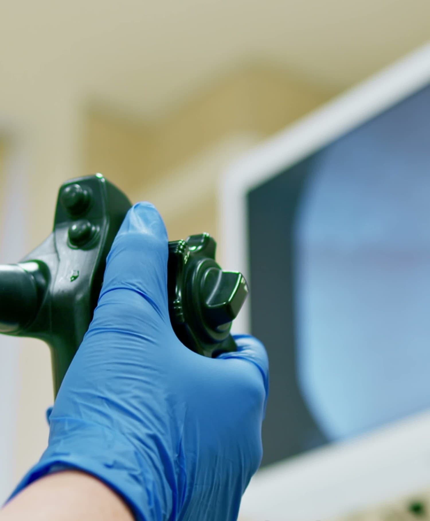 A blue-gloved hand gripping the control head of a gastroscope, the angulation dials and valves in view, with an endoscopy monitor out of focus behind.