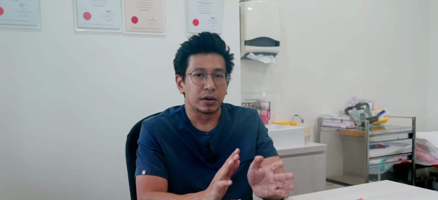 Dr Sulaiman Bin Yusof in navy scrubs at his clinic desk, gesturing as he speaks, an anatomical model of the colon on the desk beside him.