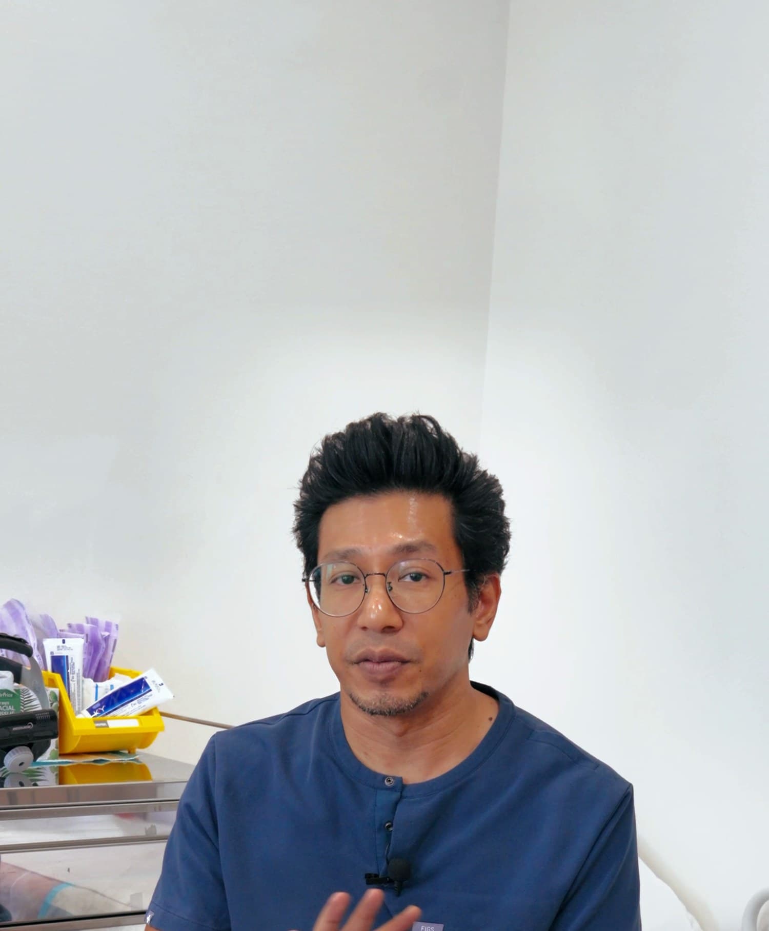 Dr Sulaiman Bin Yusof in navy scrubs seated in an endoscopy suite, gesturing with one hand as he speaks, a supply trolley with clinical consumables and the edge of a procedure bed behind him.