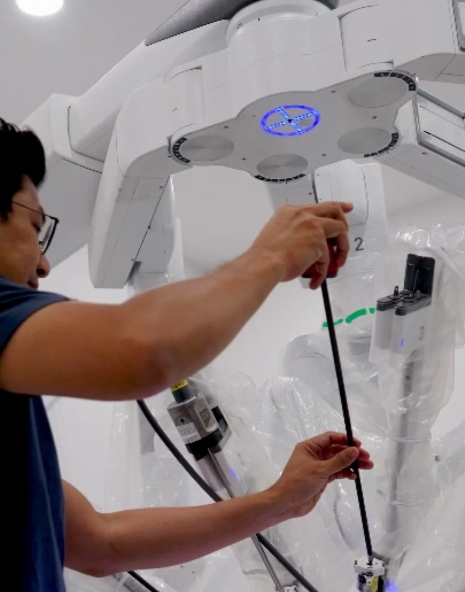 Dr Sulaiman Bin Yusof in navy scrubs reaching up to the draped arms of a da Vinci patient cart, guiding a robotic instrument down into position.