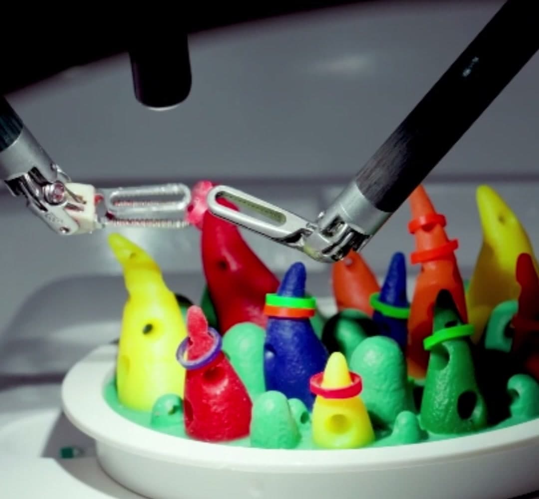 Two robotic instrument arms reaching down over a small training board of coloured rubber cones, one grasper closed around a ring it is lifting onto a peg.
