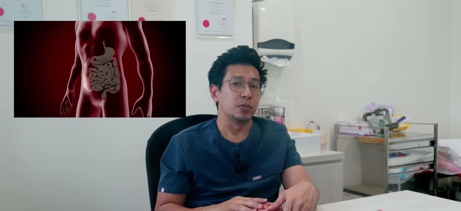 Dr Sulaiman Bin Yusof in navy scrubs, seated and speaking in a clinic room, beside an on-screen graphic of the abdomen in red showing the stomach and the intestines.