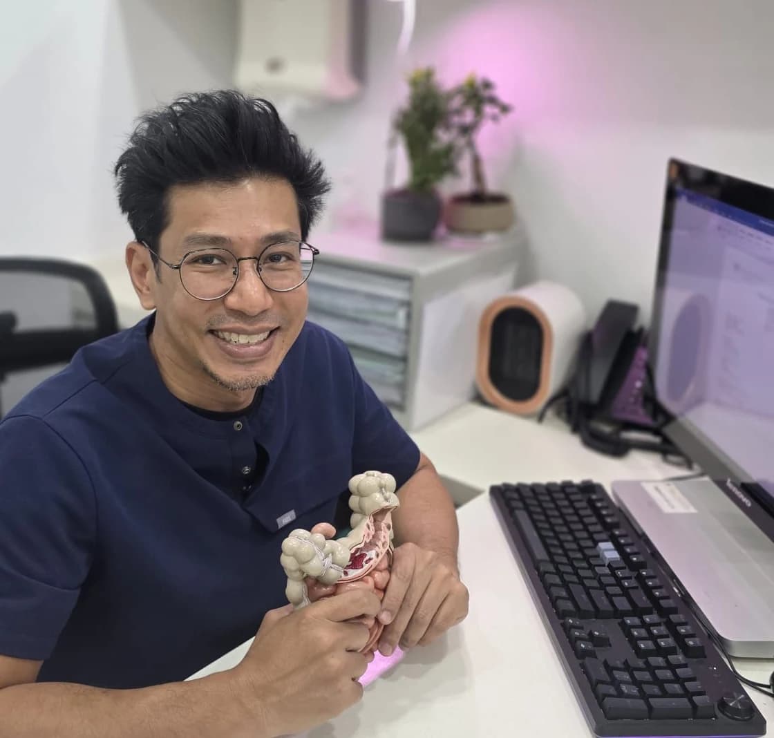 Dr Sulaiman Bin Yusof at his consultation desk in navy scrubs and round glasses, smiling and holding an anatomical model of the colon.