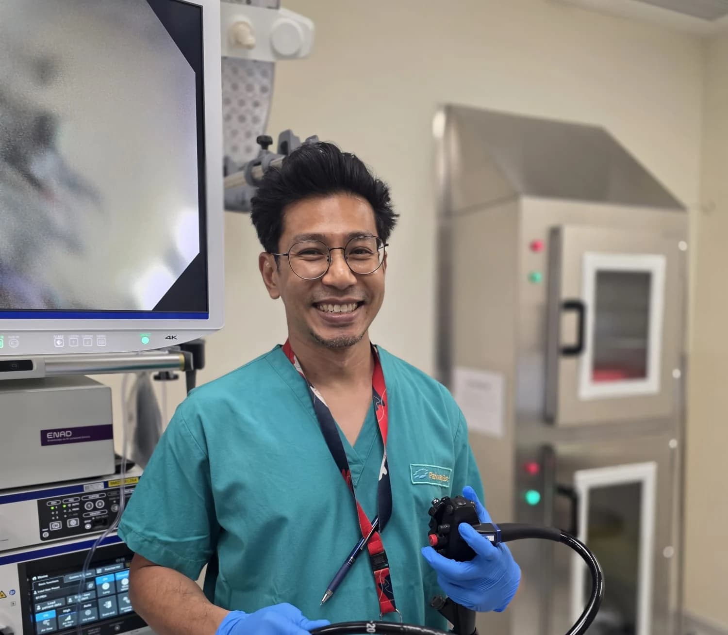 Dr Sulaiman Bin Yusof in green theatre scrubs and blue examination gloves, smiling as he holds a colonoscope in an endoscopy suite beside a 4K monitor and scope tower.
