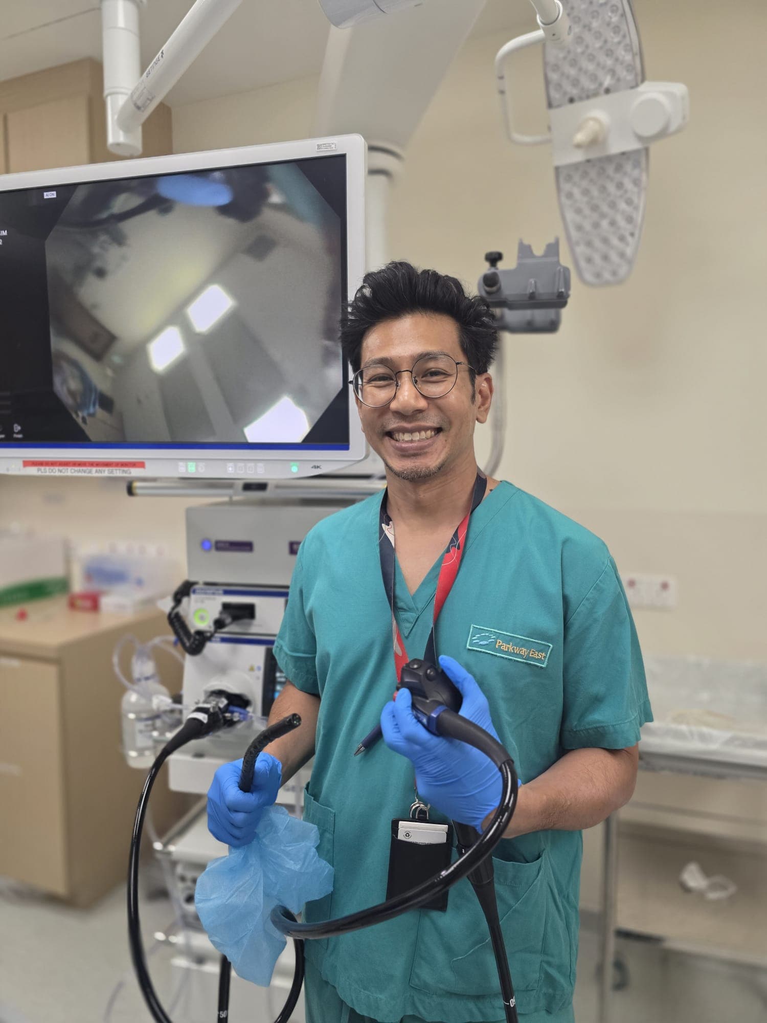 Dr Sulaiman Bin Yusof in scrubs holding an endoscope in front of the video processor stack and monitor in a hospital endoscopy suite.
