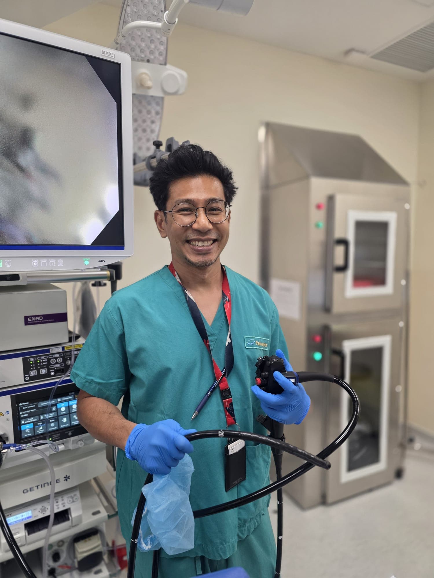 Dr Sulaiman Bin Yusof in teal surgical scrubs and blue gloves, smiling at the camera and holding a colonoscope in his endoscopy suite, with a 4K monitor to one side and the scope reprocessing cabinets behind him.