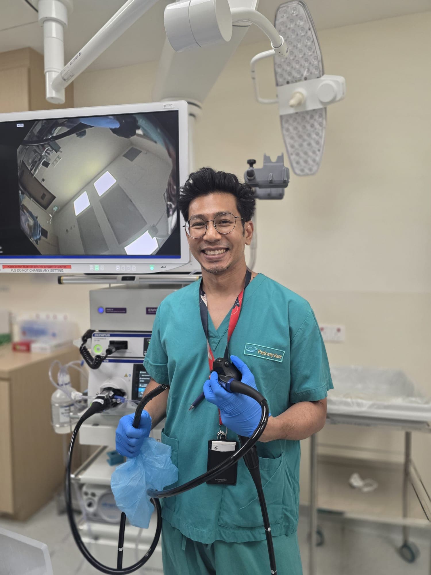 Dr Sulaiman Bin Yusof in teal surgical scrubs and blue gloves, smiling at the camera while holding an endoscope in his endoscopy suite, a 4K monitor and an overhead operating light behind him.
