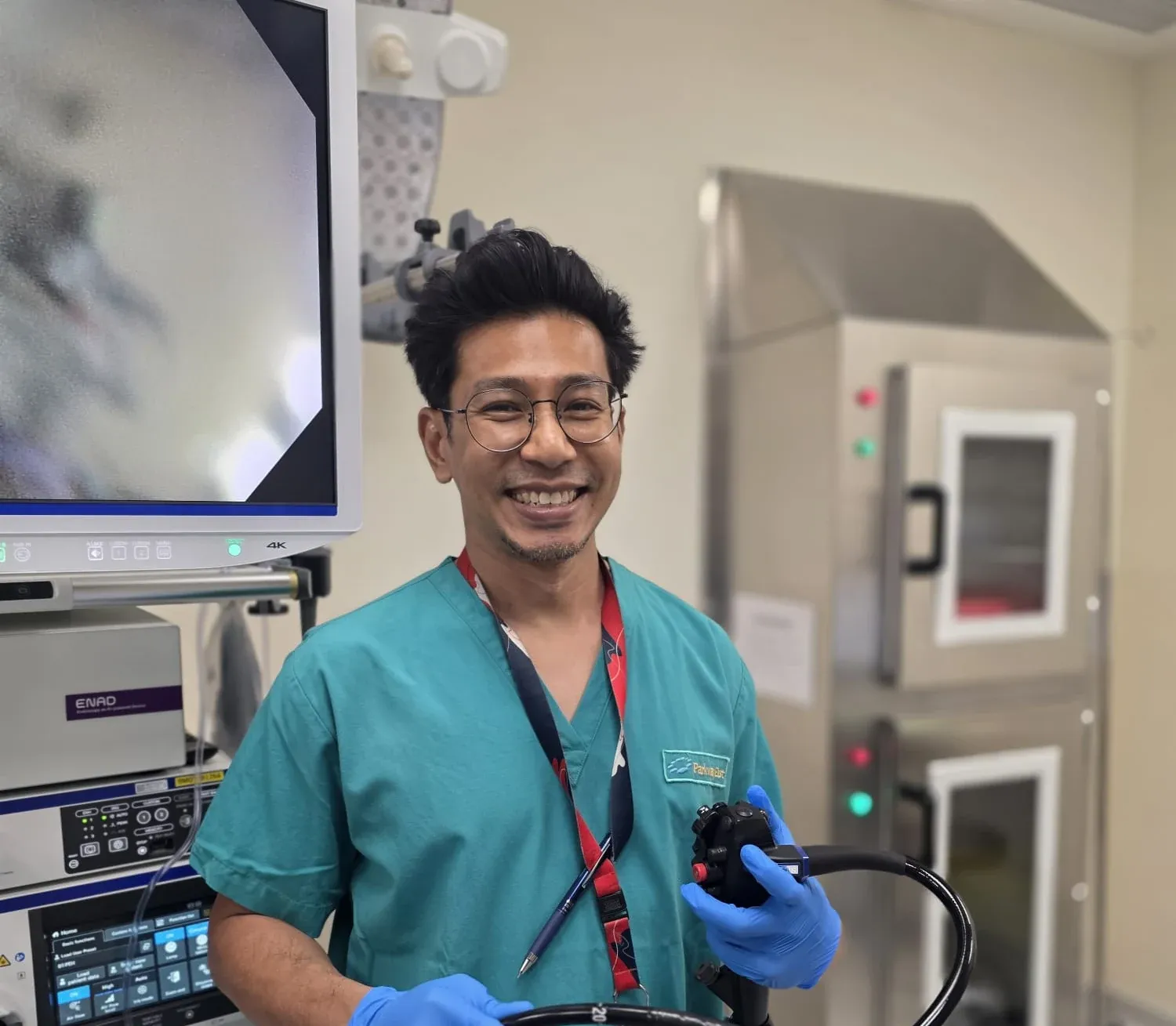 Dr Sulaiman Bin Yusof in green theatre scrubs and blue examination gloves, smiling as he holds a colonoscope in an endoscopy suite beside a 4K monitor and scope tower.